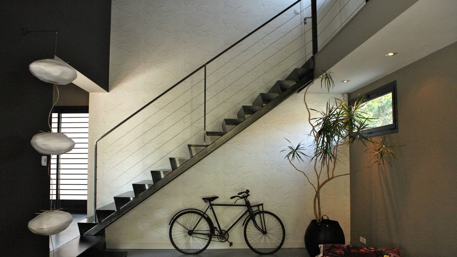 Escalier double limon central en tôle fine d’acier brut, posé sur une poutre en métal. Une rampe légère, avec une main courante en fer plat. Crédit photo : Nathalie Cousin